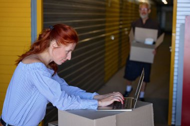 Güzel bayan bir depoda dizüstü bilgisayar kullanıyor, yanında da kutuları olan gri saçlı bir adam.