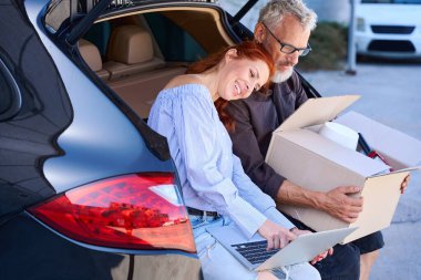 Orta yaşlı insanlar açık bagajın kenarında otururken, bir kadın başını kocasının omzuna yasladı.