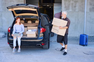 Kadın elinde dizüstü bilgisayarla bagajın kenarına oturmuş, elinde karton kutular olan bir adamın yanında.