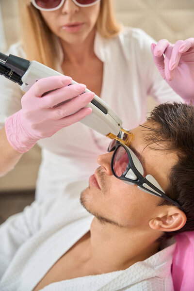 Young client of a beauty salon undergoes a laser facial hair removal procedure, a specialist uses modern equipment