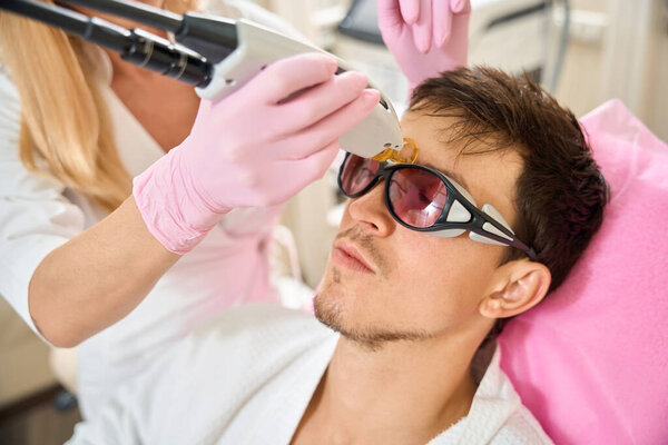 Beauty salon client undergoing laser facial hair removal procedure, specialist uses modern equipment