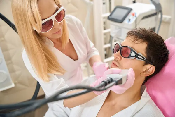 Blonde woman performs laser hair removal procedure on a young mans face, people wearing safety glasses