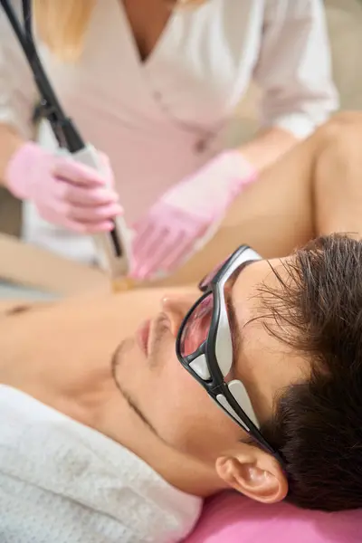 Client of a cosmetology salon during a laser hair removal session in the armpit area, a man wearing safety glasses