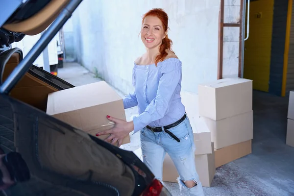Smiling woman took a cardboard box from the trunk of a car, she is in casual clothes