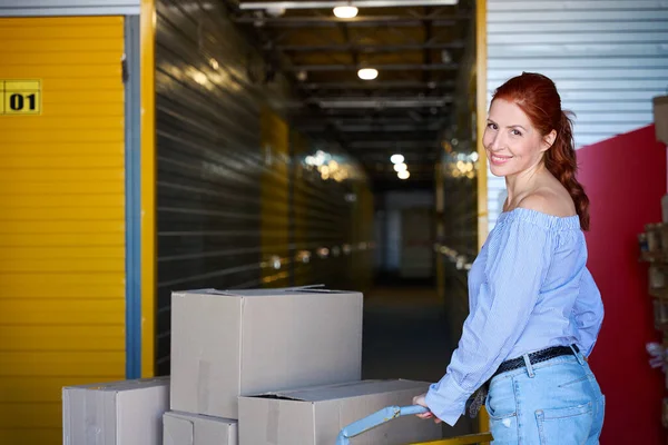 Smiling female is pushing a cargo cart through a warehouse, there are a lot of cardboard boxes on the cart