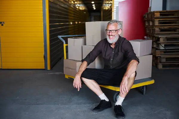 Cheerful man sat down to rest on a cart with boxes, he is in a warehouse