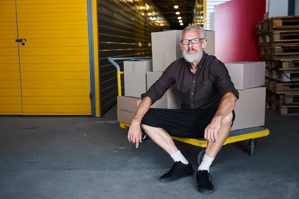 Man with glasses sat down to rest on a cart with boxes, he is in a warehouse