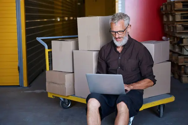 Middle-aged man sat down on a cart with boxes, he communicates on a laptop