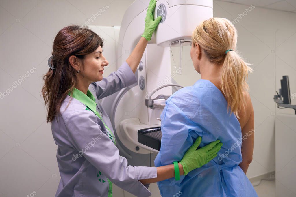 Radiologic technologist operating control panel on mammography machine for positioning compression paddle against patient chest wall