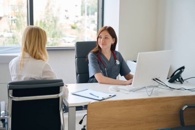 Muayenehanesinde oturan hekim masaüstü bilgisayarında müşteriye bakarken daktilo yazıyor.