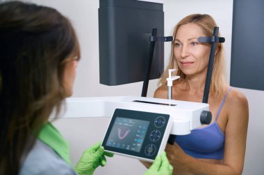 Experienced radiologic technologist positioning control panel and bite block in front of patient mouth before dental radiography clipart