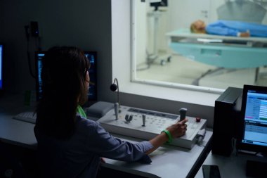 Kontrol odasının penceresinden hastayı röntgen masasında izlerken radyo teknolojisi uzmanının kumanda panelinde joystick kullanışının arka görüntüsü