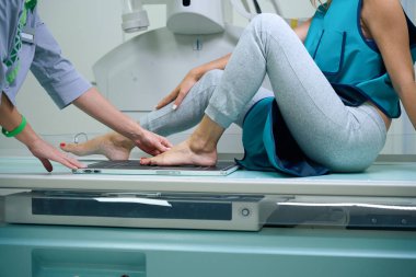 Radyolojik teknoloji uzmanı ayağını plantar yüzeyiyle görüntüleme reseptörüne koyarken radyografik masada oturan bir kadının kesilmiş fotoğrafı.