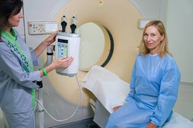 Deneyimli radyolojik teknoloji uzmanı yetişkin kadını kontrast medyayla bilgisayarlı tomografiye hazırlıyor.