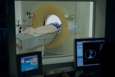 Tomografi odasındaki muayene masasında sırtüstü uzanan yetişkin bir kadın kontrol odasındaki radyograf tarafından izleniyor.