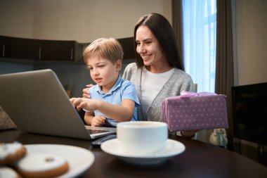 Neşeli bir kadın, oğluyla dizüstü bilgisayarın önünde oturmuş kış tatilini kutlayan bir video görüşmesi yapıyor.