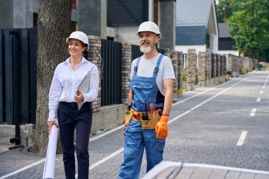 Joyful site manager and builder standing on street in front of unfinished residential house clipart