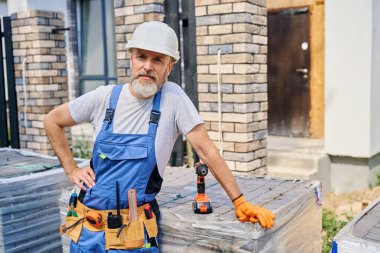 Güvenlik miğferi takmış inşaatçı bitmemiş bir evin önündeki beton kaldırım bloklarına yaslanıyor.