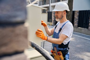 Serious electrician in safety helmet tightening screws on power switchboard with screwdriver clipart