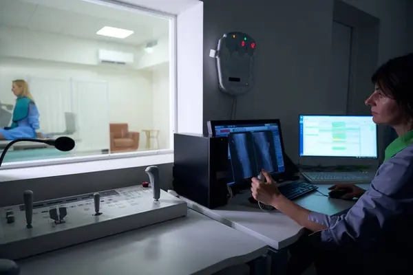 Stock image Focused radiographer seated at desktop computers looking at woman on radiographic table through window in control room