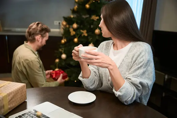 漂亮的女人在家里的圣诞树边喝咖啡 男人拿着礼物 — 图库照片