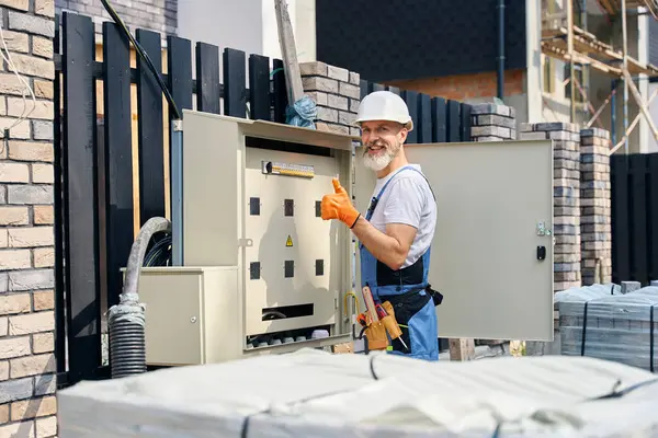 Joyful electrical engineer in safety helmet giving thumbs-up while standing outdoors in front of open power distribution box