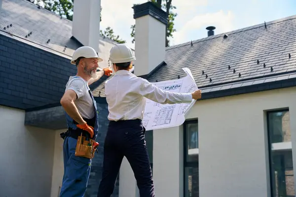 建物の点検の間に場所のスーパーバイザーに新しい家の屋根を指す笑顔の建築者 — ストック写真