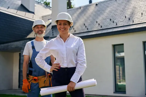新築住宅の前に立つ笑顔の女性サイトスーパーバイザーと喜ばしい建築請負業者 — ストック写真