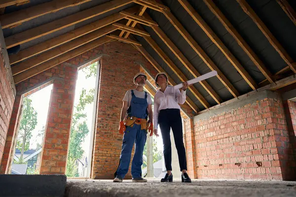 未完成の住宅ビルの内部からの屋根を点検する建築図面および請負業者が付いている集中されたプロジェクト マネージャー — ストック写真