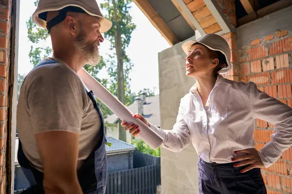 Irate female construction superintendent with blueprints in hand arguing with contractor inside unfinished residential house