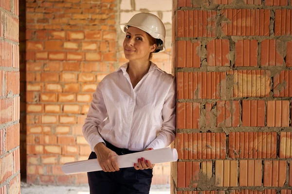 Zadowolona Inspektorka Budowlana Planami Opartymi Ścianę Cegły Pół Wybudowanym Domu — Zdjęcie stockowe