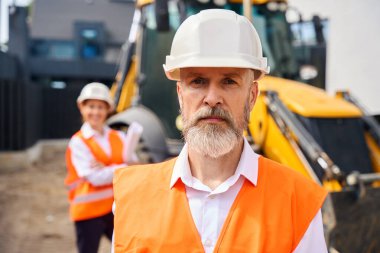 Ciddi bir ustabaşı arka planda kadın mühendis ile kepçenin önünde duruyor.