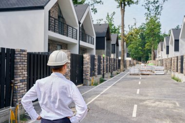 Asfalt caddenin ortasında duran inşaat müdürünün arka görünümü özel konut evlerinin satırları arasında