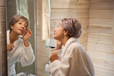 Bornozlu emekli, aynadaki yansımasına hayran. Banyo temiz ve rahat.