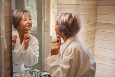 Bornozlu kadın aynadaki yansımasına hayran. Banyoda bir sürü bakım ürünü var.