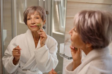 Bornozlu yaşlı kadın yeşim bir yüz masajı aleti kullanıyor, banyo temiz ve rahat.