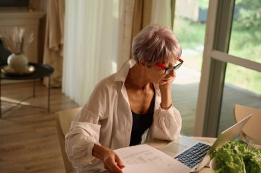 Evde, mutfakta beyaz tişörtlü bir kadın, dizüstü bilgisayarı ve çalışma kağıtları, masada taze sebzeler.