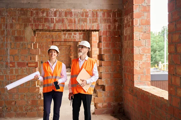 Smiling construction manager and serious contractor standing in room of half-built private dwelling house