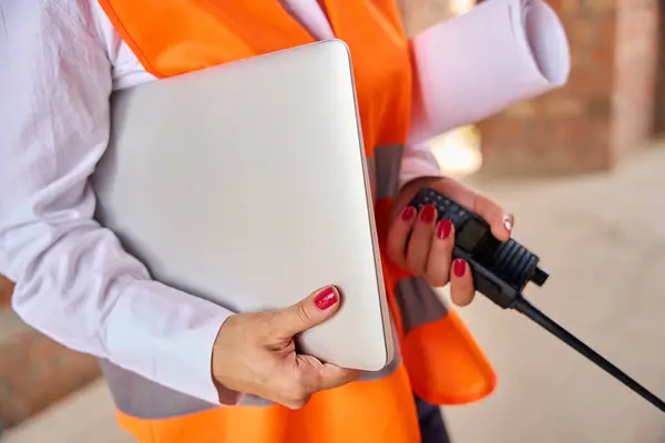 Cropped photo of construction supervisor with rolled-up blueprints under arm holding walkie-talkie and laptop while standing in unfinished dwelling house