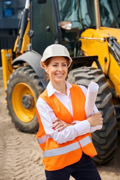 Smiling building inspector with rolled-up architectural drawings under arm looking into distance while standing near backhoe loader