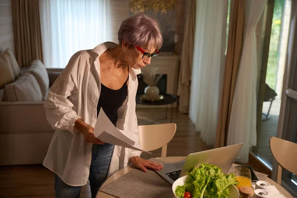 Lady with glasses at home in the kitchen with a laptop and work papers, fresh vegetables on the table