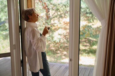 Bayan pencerenin dışındaki doğaya bakarken bir fincan kahveden hoşlanır.