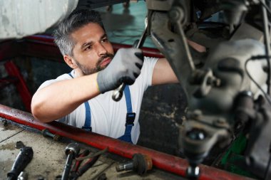 Serious car service technician is tightening nut on automobile underbody with wrench clipart