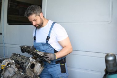 Odaklanmış oto servis teknisyeni iş kıyafetleri içinde garajda minibüsün yanında dururken silindir kafa örtüsünü açıyor