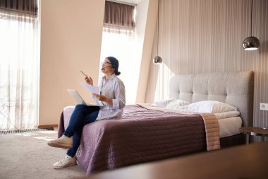 Otel yatağında elinde bilgisayar ve belgelerle oturmuş internet telefon görüşmesi sırasında mikrofona konuşan bir kadın girişimci.