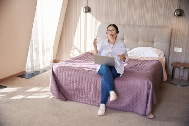 İş kadını, otel odasındaki internet görüşmesi sırasında bilgisayar ve belgelerle birlikte mikrofona oturmuş konuşuyordu.