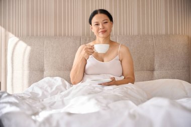 Elinde bir bardak içecekle ve elinde fincan tabağıyla otel odasında oturan memnun bir bayan.