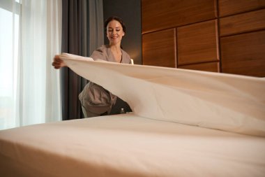 Üniforma giymiş gülümseyen oda hizmetçisi otel odasındaki yatağa temiz battaniye seriyor.
