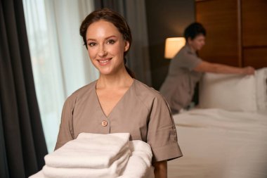 Meslektaşı misafir odasında yatağını hazırlarken elinde bir yığın temiz havluyla kameraya poz veren neşeli hizmetçi.