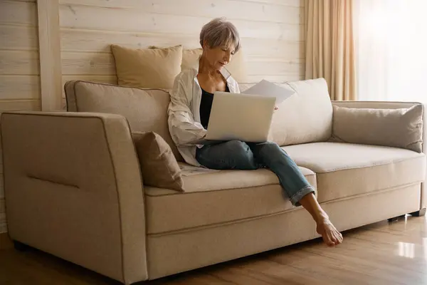 Elderly woman with short haircut sits with a laptop and work papers on a cozy sofa in an eco-design room